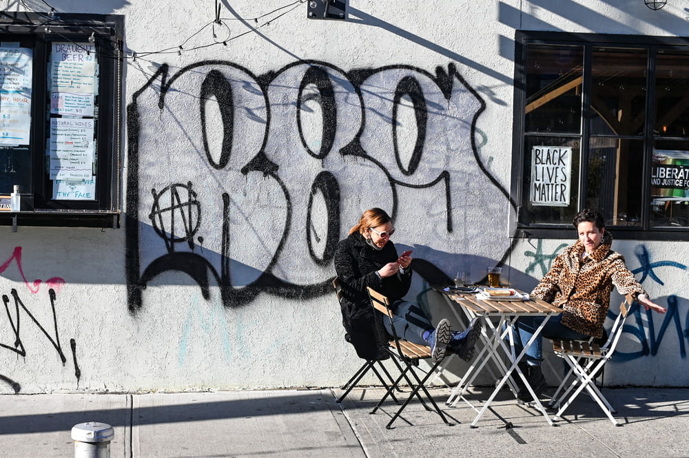 man in black jacket sitting on folding chair beside wall with graffiti during daytime
