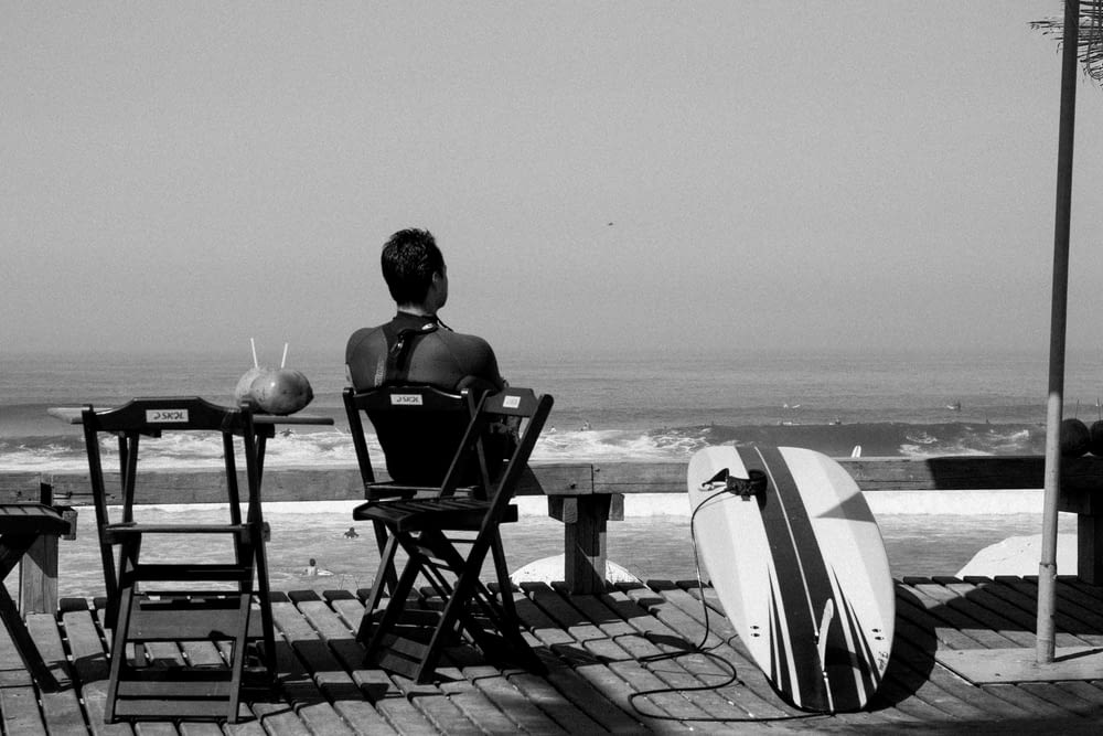 grayscale photo of woman sitting on chair near body of water