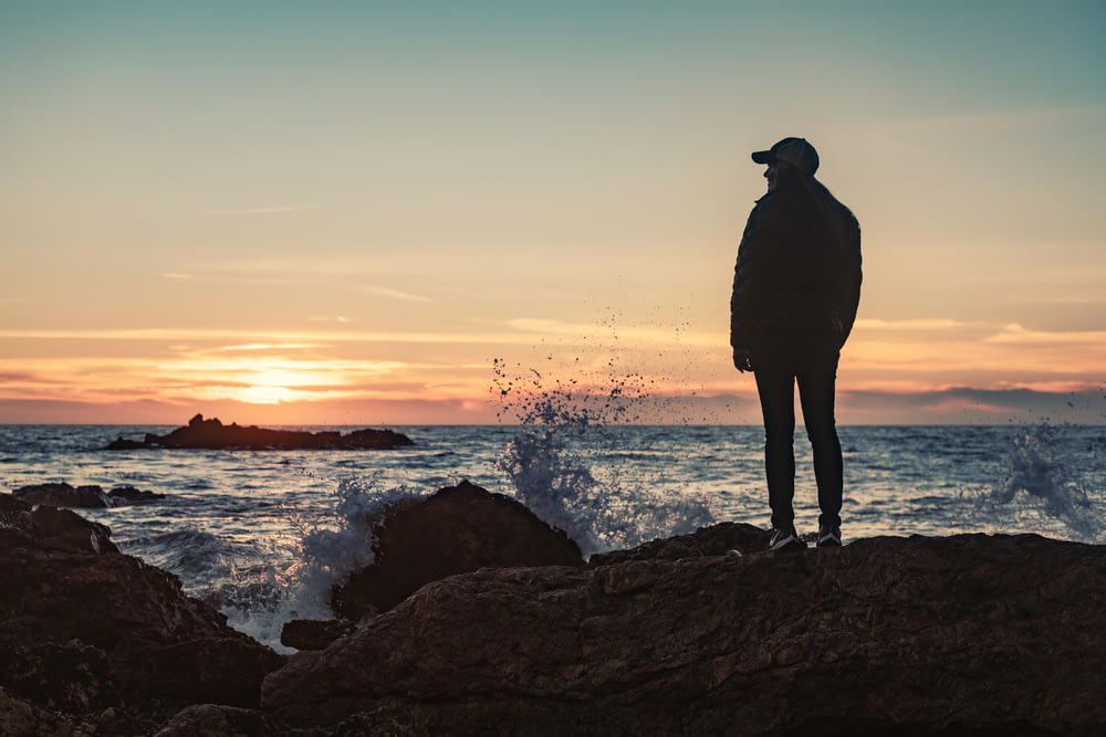 Silhouette d’homme debout sur la formation rocheuse près du plan d’eau au coucher du soleil