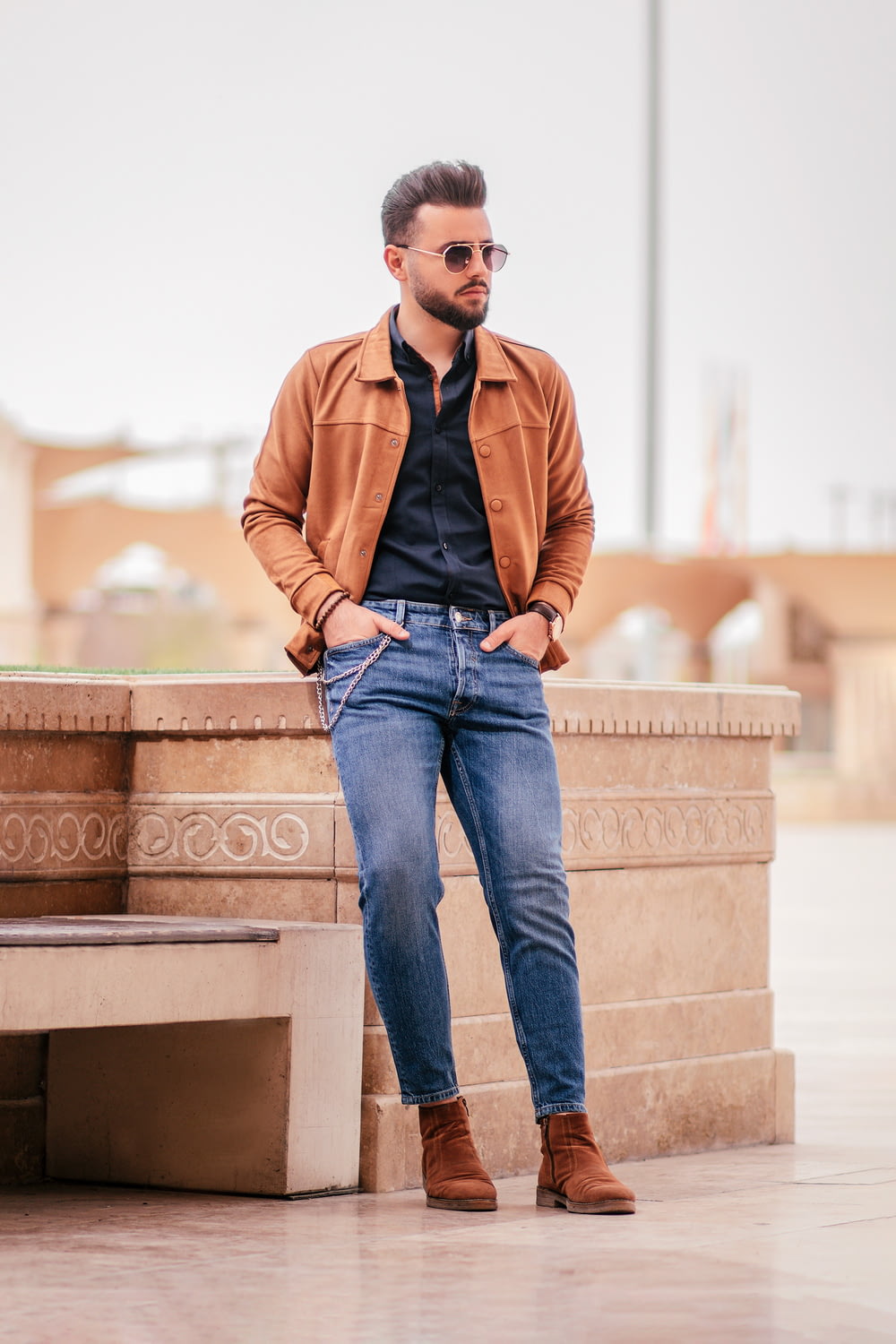 Hombre con chaqueta marrón y jeans de mezclilla azul sentado en un banco de concreto marrón durante el día