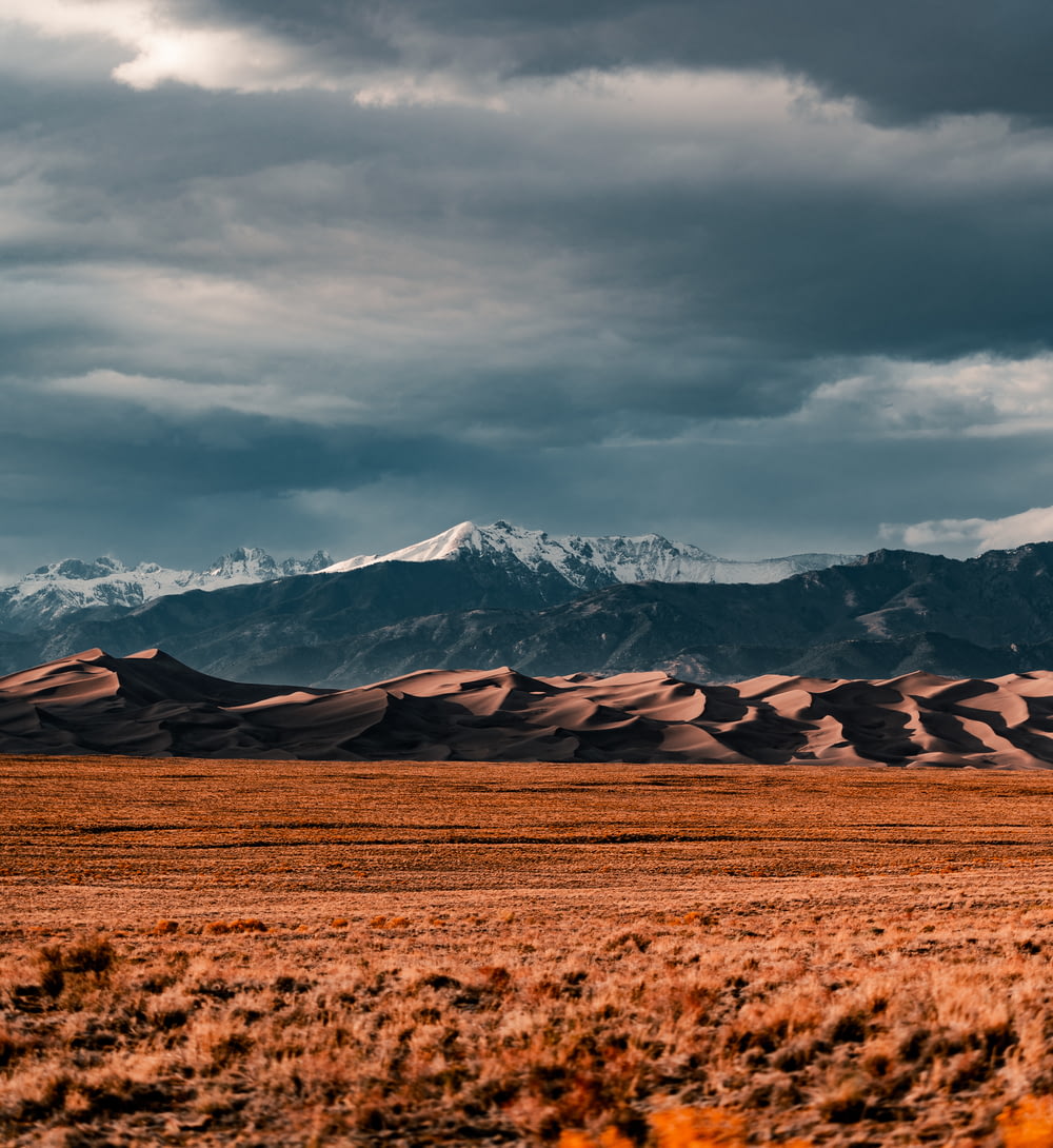 brown field near snow covered mountains under cloudy sky during daytime