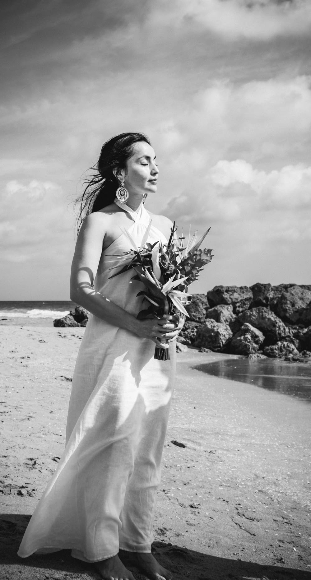 woman in white spaghetti strap dress holding bouquet of flowers standing on beach shore