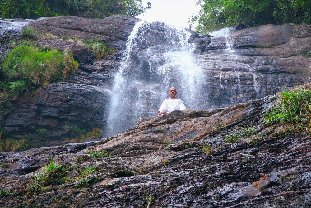 uomo in camicia bianca che si siede sulla roccia marrone vicino alle cascate durante il giorno