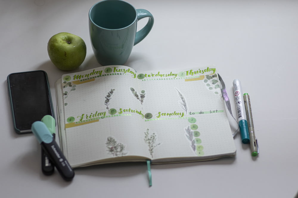 Taza de cerámica verde junto a bolígrafo de clic negro en libro blanco y verde