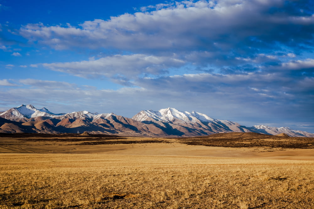 campo marrom perto de montanhas cobertas de neve sob céu nublado azul e branco durante o dia