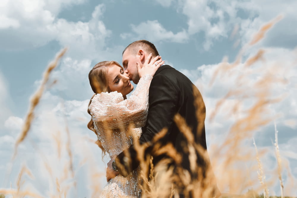 man in black suit kissing woman in white lace dress under blue and white cloudy sky