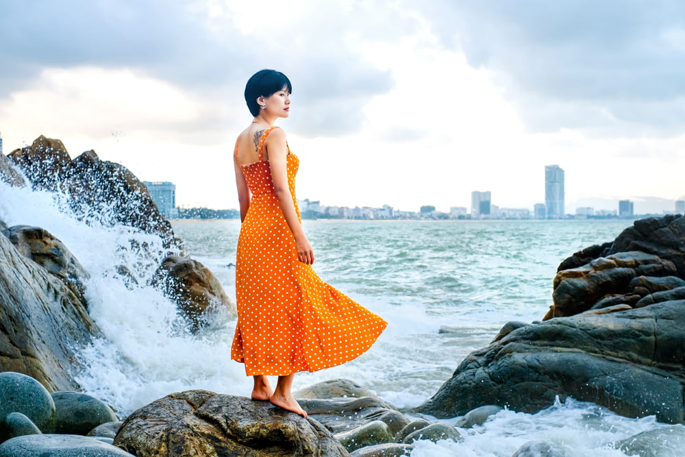 woman in red and white polka dot dress standing on rock near sea during daytime