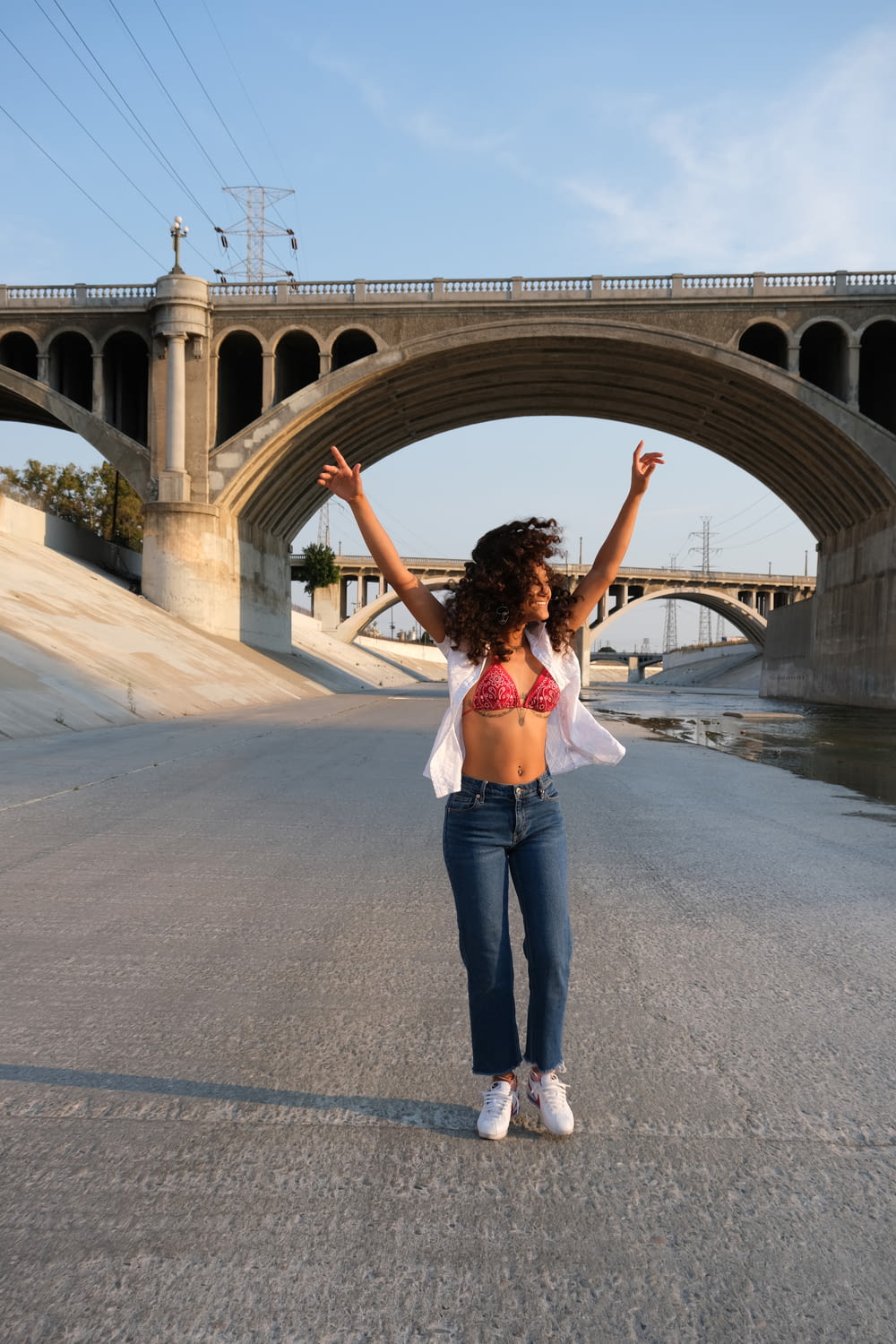 Donna in crop top bianco e jeans blu in denim in piedi sul ponte di cemento grigio durante il giorno