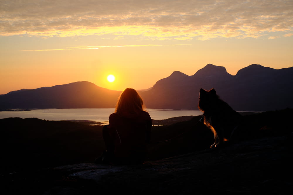 a woman and a dog sitting on a hill watching the sun go down