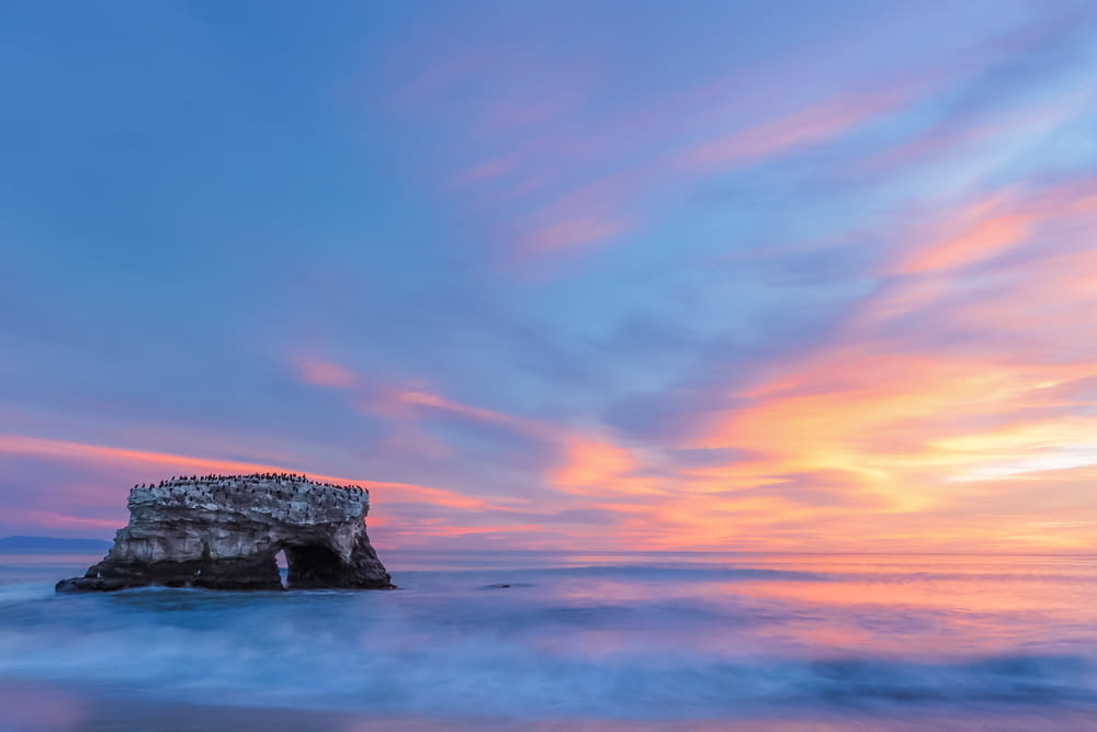 a large rock sticking out of the ocean under a colorful sky