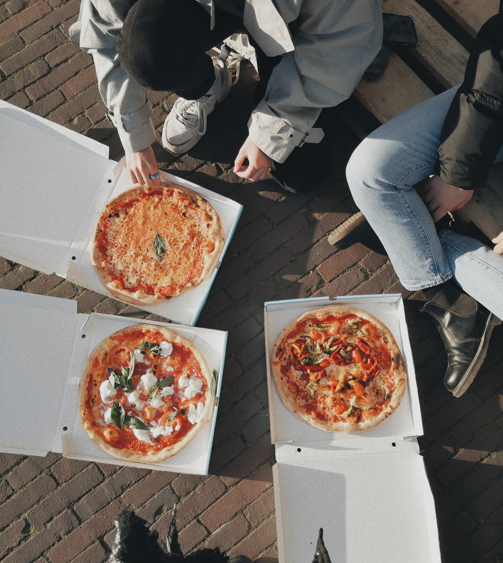 three people sitting on a bench with three pizzas in boxes