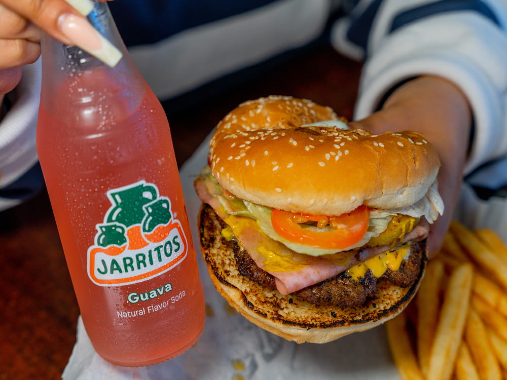 a person holding a large hamburger next to a bottle of juice