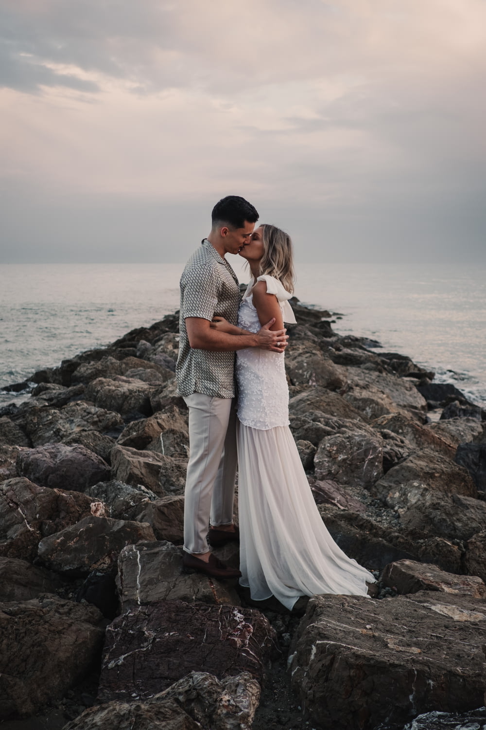 a bride and groom kissing on the rocks by the ocean