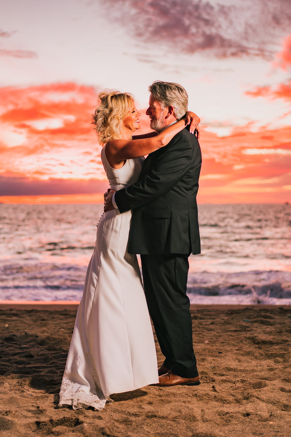 a bride and groom embracing on the beach at sunset