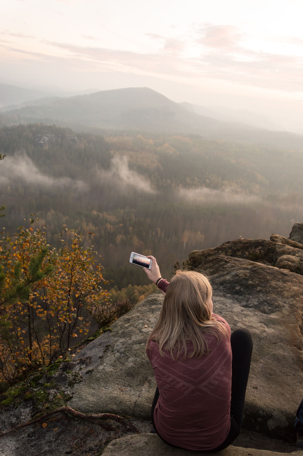 a woman sitting on top of a cliff taking a picture