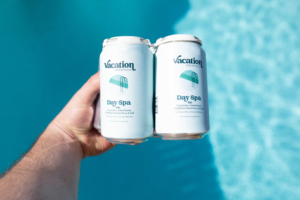 a hand holding two cans of vacation by the pool