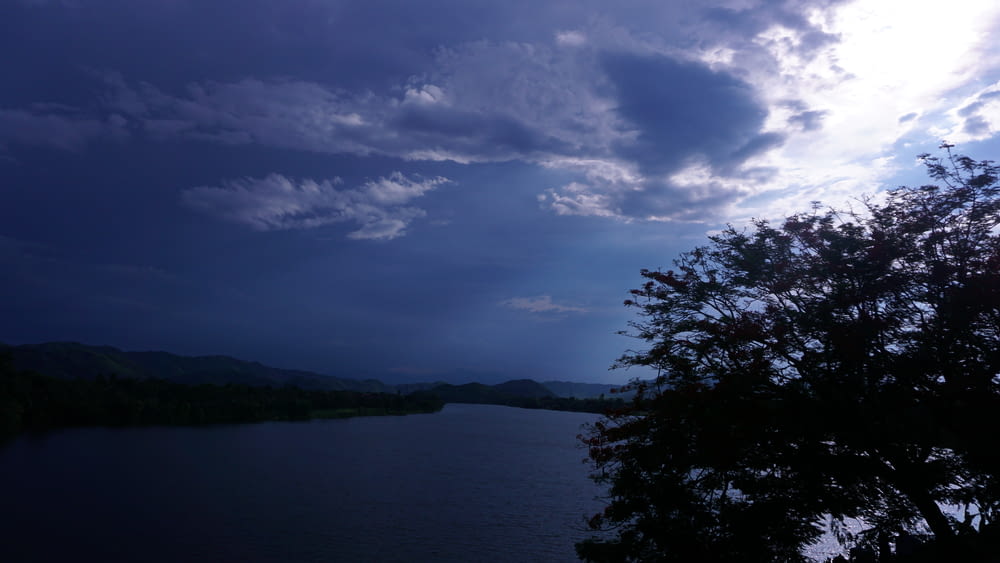 a lake with a tree in the foreground and clouds in the background