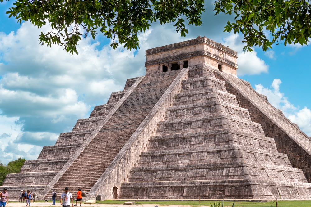 a large pyramid with people standing around with Chichen Itza in the background