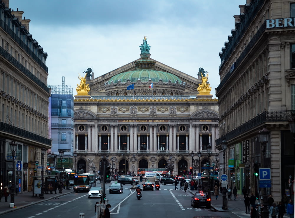 a street with cars and buildings on either side of it with Palais Garnier in the background