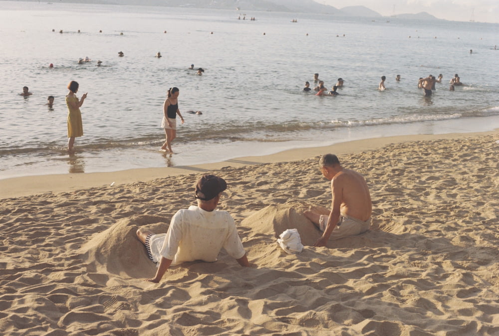 a couple of men sitting on a beach with people in the water