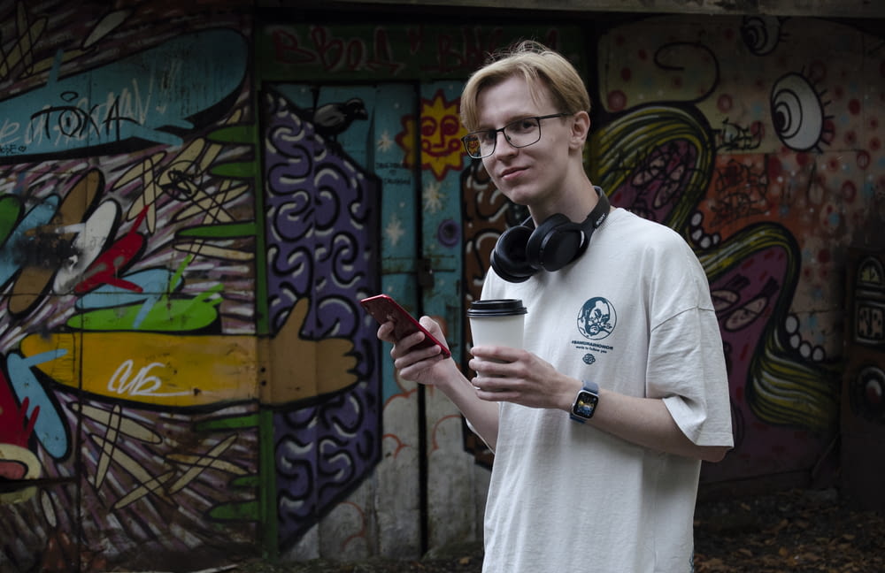 a man wearing headphones and holding a cup in front of a wall with graffiti