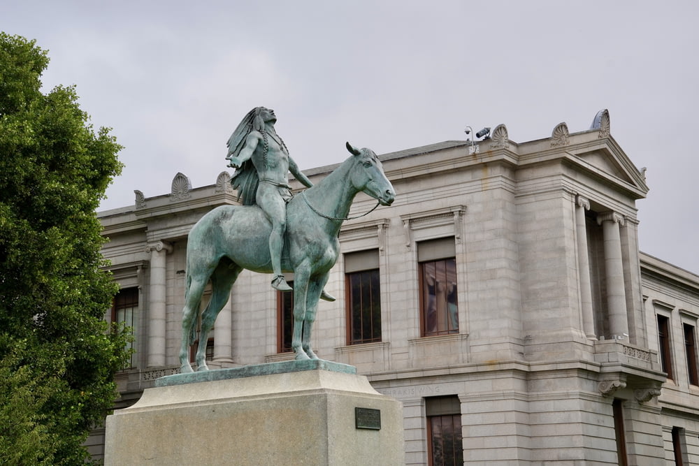 eine Statue einer Person, die vor einem Gebäude auf einem Pferd reitet