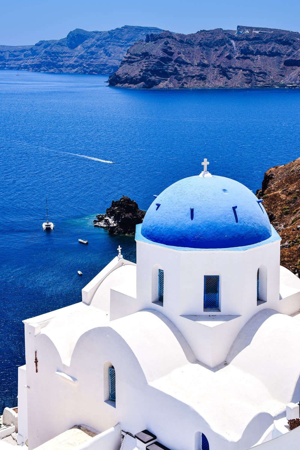a white building with a blue roof by a body of water with Santorini in the background