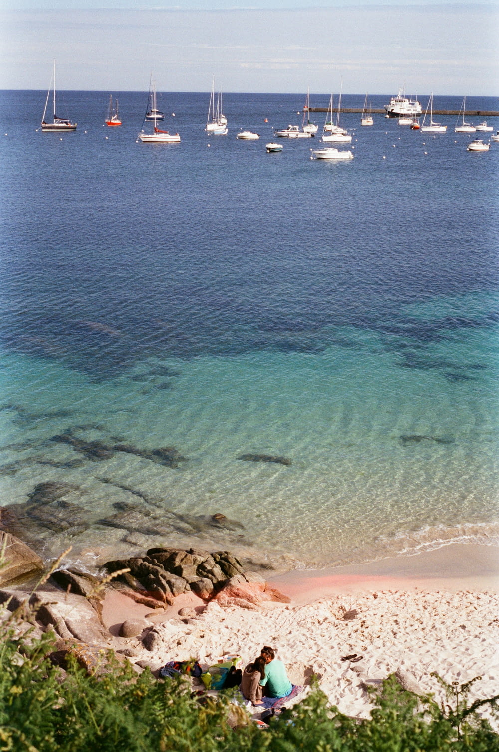 a group of people sitting on a rocky beach with boats in the water