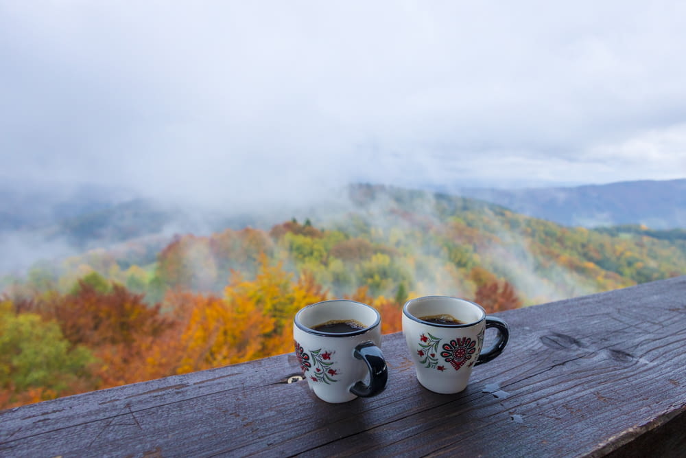 two mugs of coffee sit on a wooden ledge overlooking a mountain