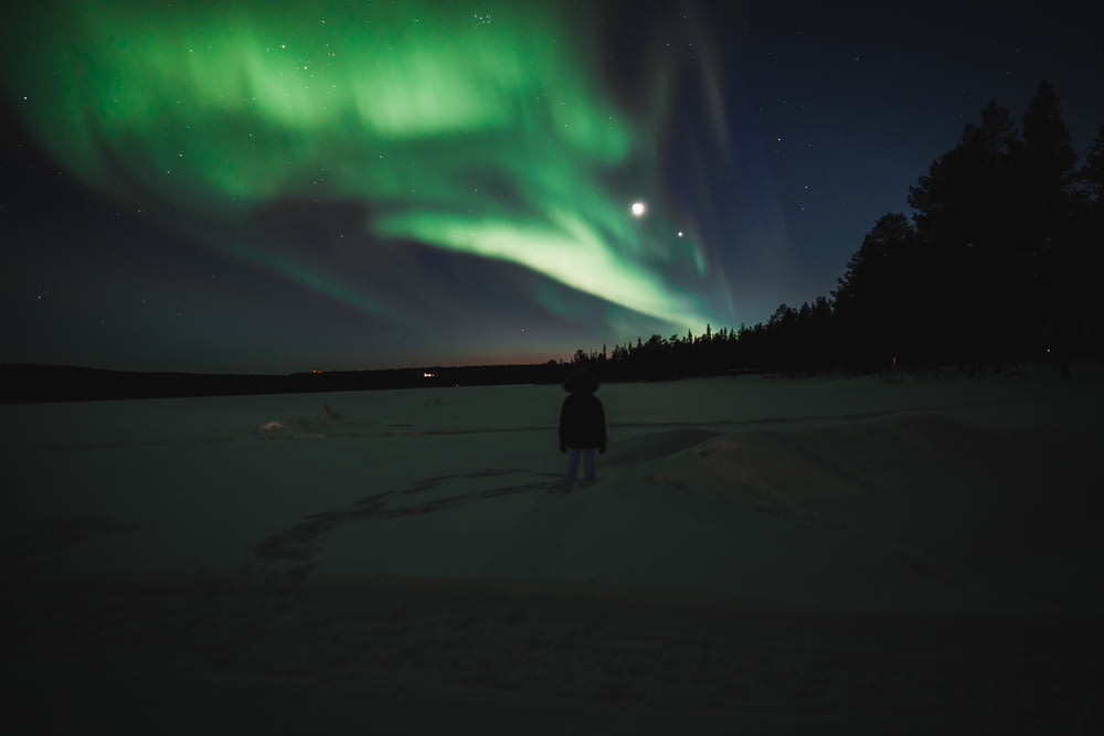 a person standing in the snow under a green aurora bore