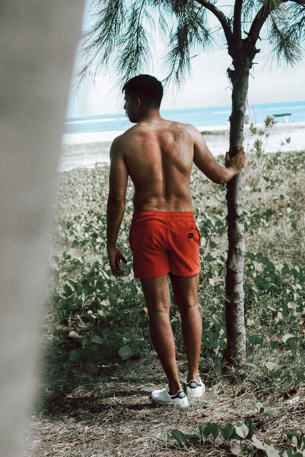 ein Mann ohne Hemd, der an einem Strand neben einem Baum spazieren geht