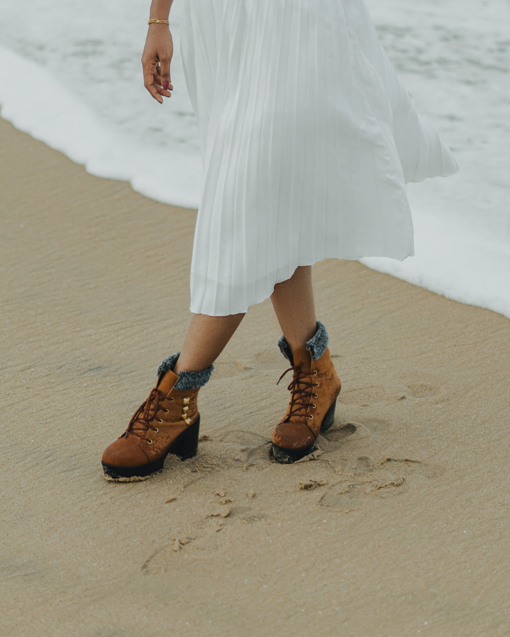 a woman in a white dress and brown boots walking on the beach