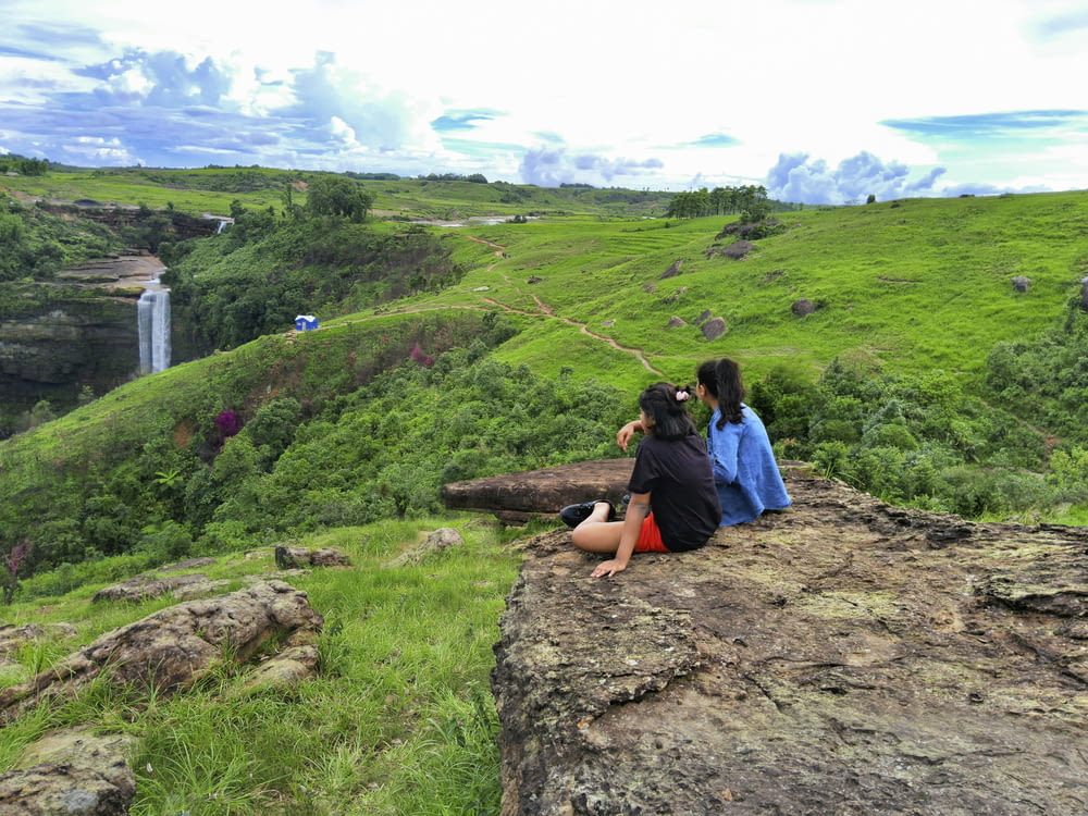 two people sitting on a rock looking at a waterfall