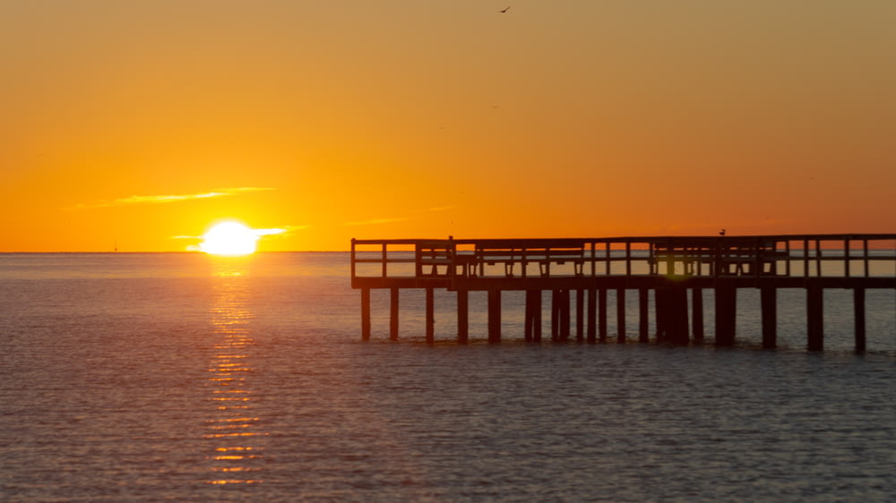 the sun is setting over the ocean with a pier in the foreground