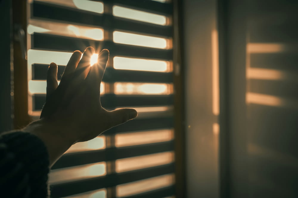 a person's hand reaching for the light coming through the blinds