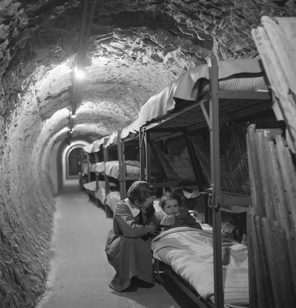 Woman kneeling next to bed of child, in an underground tunnel during the bombing of London during World War II
