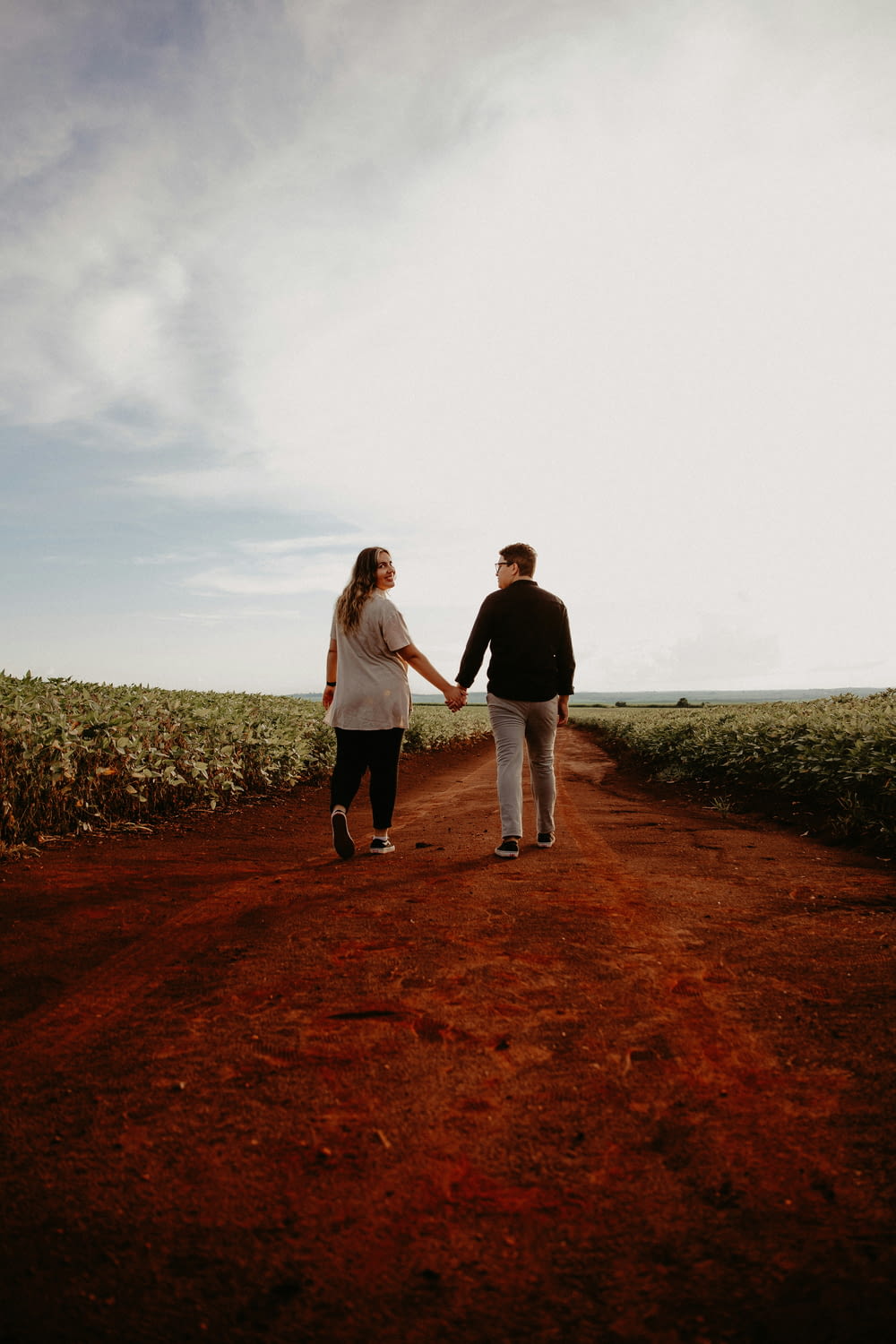 a man and woman holding hands walking down a dirt road