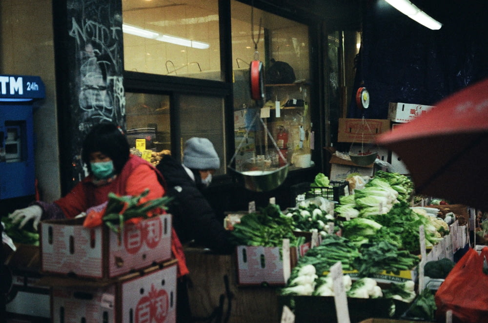 a woman wearing a face mask standing in front of a vegetable stand