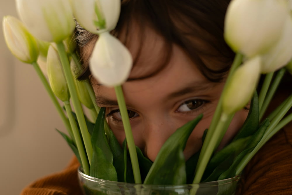 a girl hiding her face behind a vase of white tulips