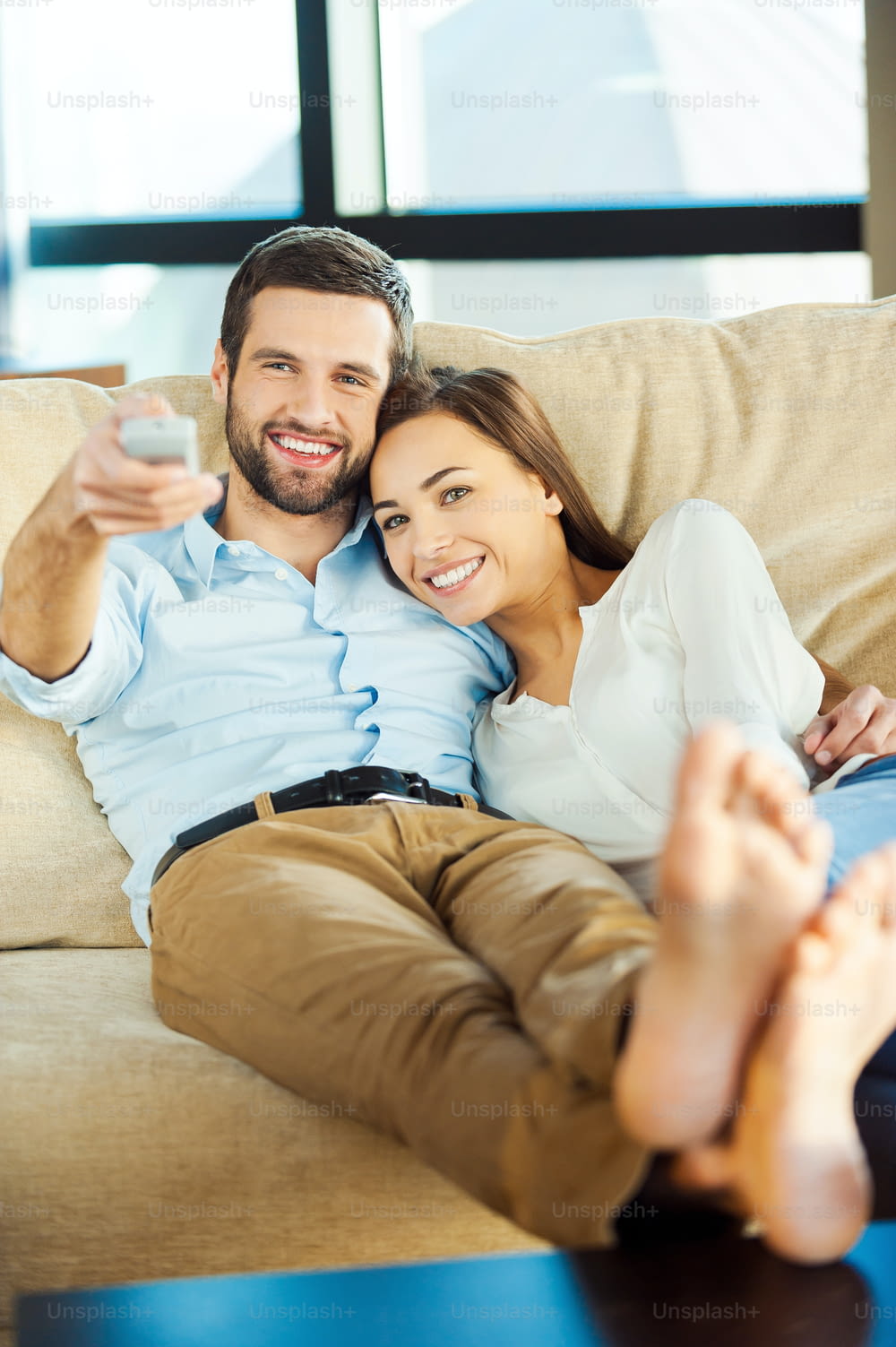 아름다운 젊은 사랑하는 커플이 소파에 함께 앉아 TV를 보고 있는 동안 남자는 리모컨을 들고 웃고 있다