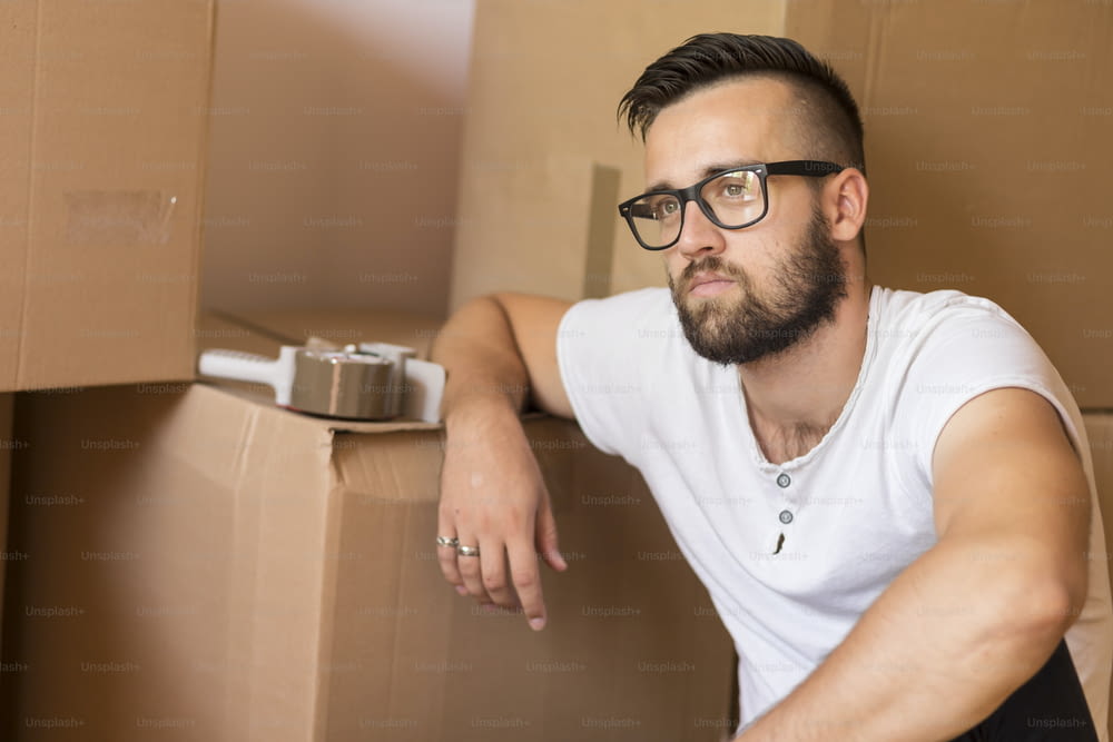 Young man moving in a new apartment, sitting on the floor, surrounded with cardboard boxes with packing machine placed on one of the boxes in the background