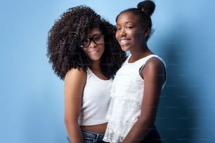 カメラを見ながら微笑む2人の美しいアフリカ系アメリカ人の女の子。青い背景にポーズをとる姉妹。スタジオ撮影。