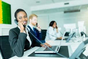Une belle jeune femme noire travaillant dans un centre d’appels dans un bureau avec son partenaire aux cheveux roux à l’autre bout du bureau en train de parler à un autre client