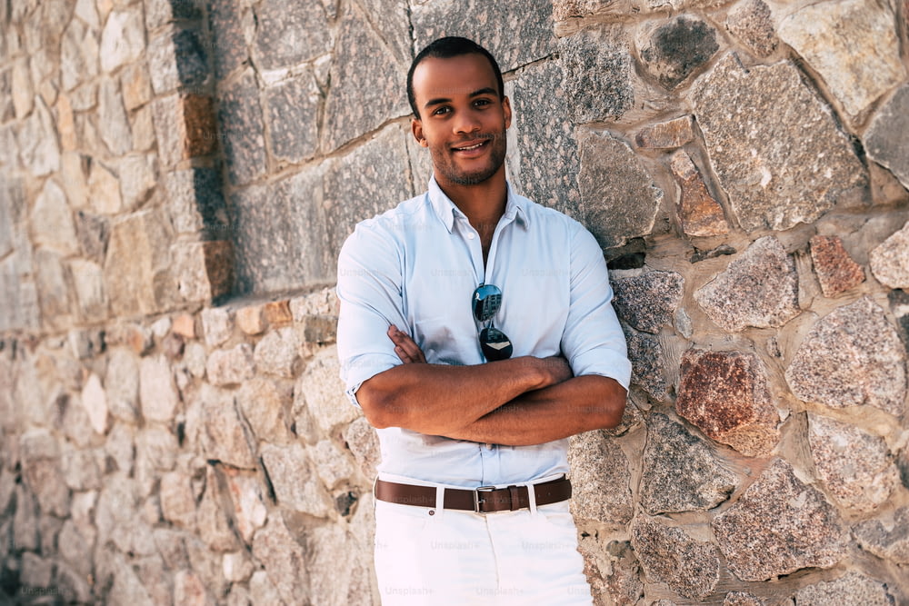Handsome young African man in smart casual clothes keeping arms crossed and looking at camera with smile while standing against the stoned wall outdoors