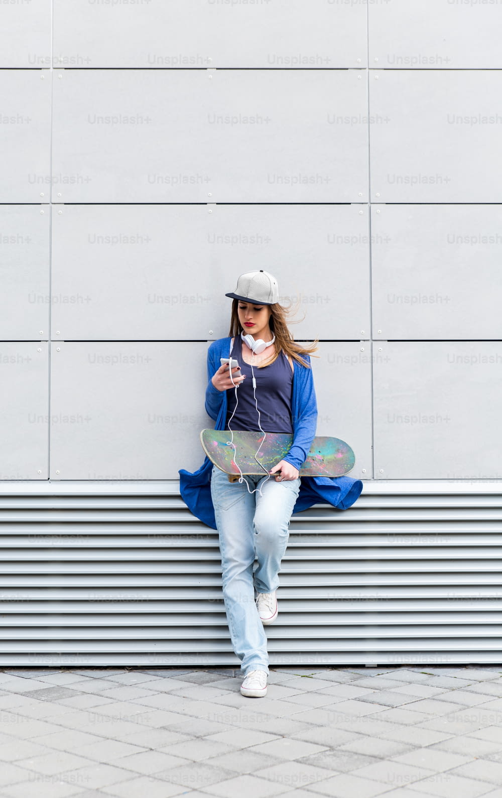 Young girl with skateboard leaning on modern gray wall, she is holding a mobile phone in her hand.