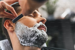Nahaufnahme von Kosmetikerhänden, die Rasiermesser zum Rasieren des männlichen Bartes verwenden. Junger Mann schloss die Augen vor Entspannung