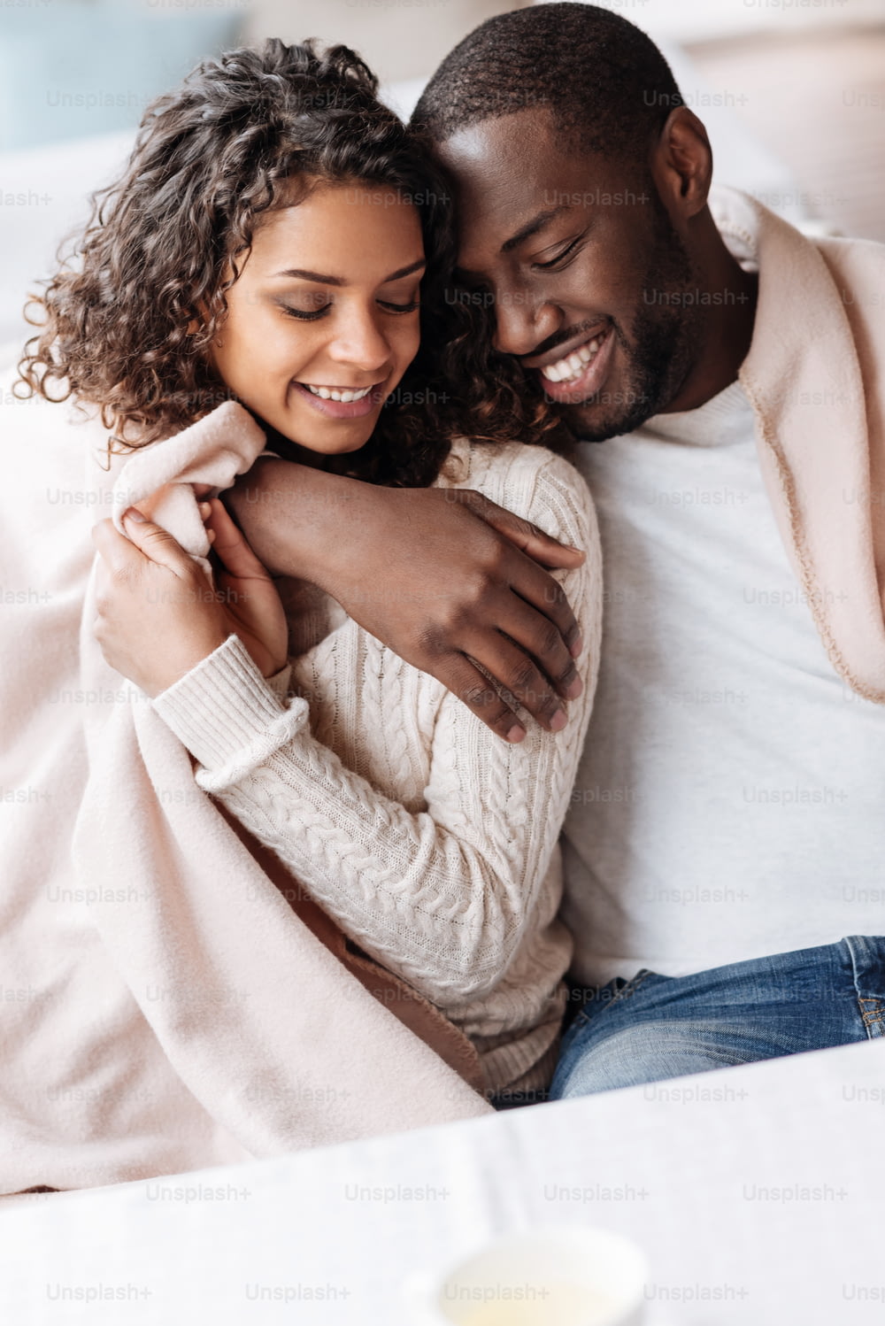 Happy to be in love. Positive young delighted African American couple sitting in the cafe and being covered with a blanket while expressing love and hugging