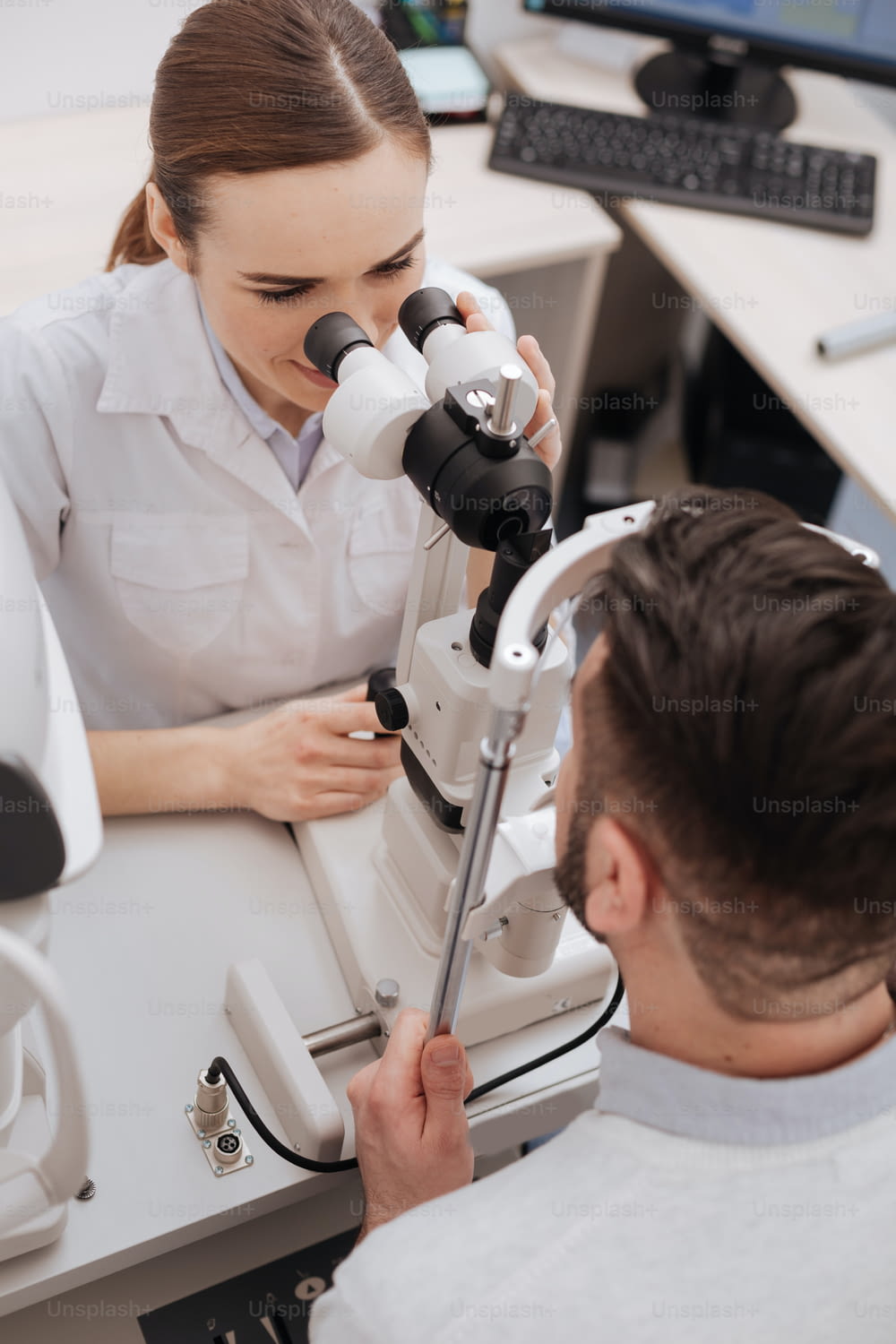 Ophthalmologische Untersuchung. Draufsicht einer netten professionellen Optikerin, die ihrem Patienten gegenüber sitzt und seine Augen untersucht, während er medizinische Geräte benutzt