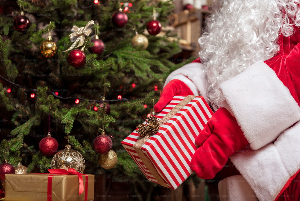 Close up of Santa Claus hands laying Christmas gift under the fir-tree. He is wearing red gloves