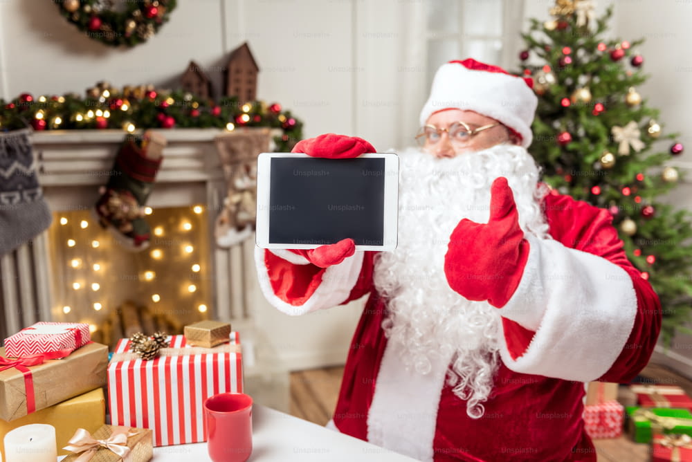 Le meilleur cadeau de tous les temps. Un vieil homme barbu joyeux en costume présente une tablette et lève le pouce. Il est assis à table près des boîtes de cadeaux de Noël. Concentrez-vous sur le gadget